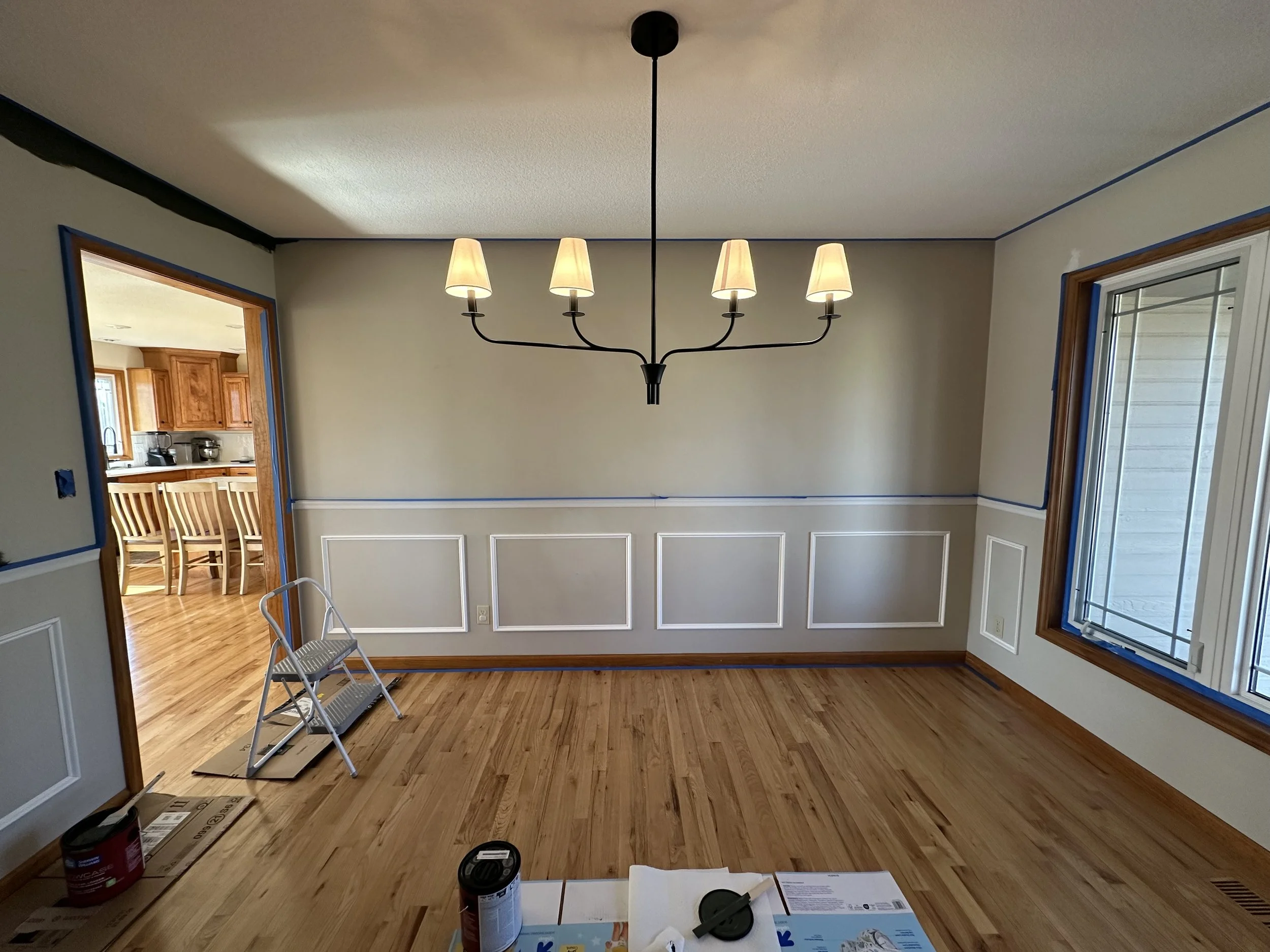 Empty dining room with wooden flooring, a large window on the right, and a chandelier hanging from the ceiling. Painting supplies and a small step ladder are on the floor, indicating ongoing decorating or painting work.