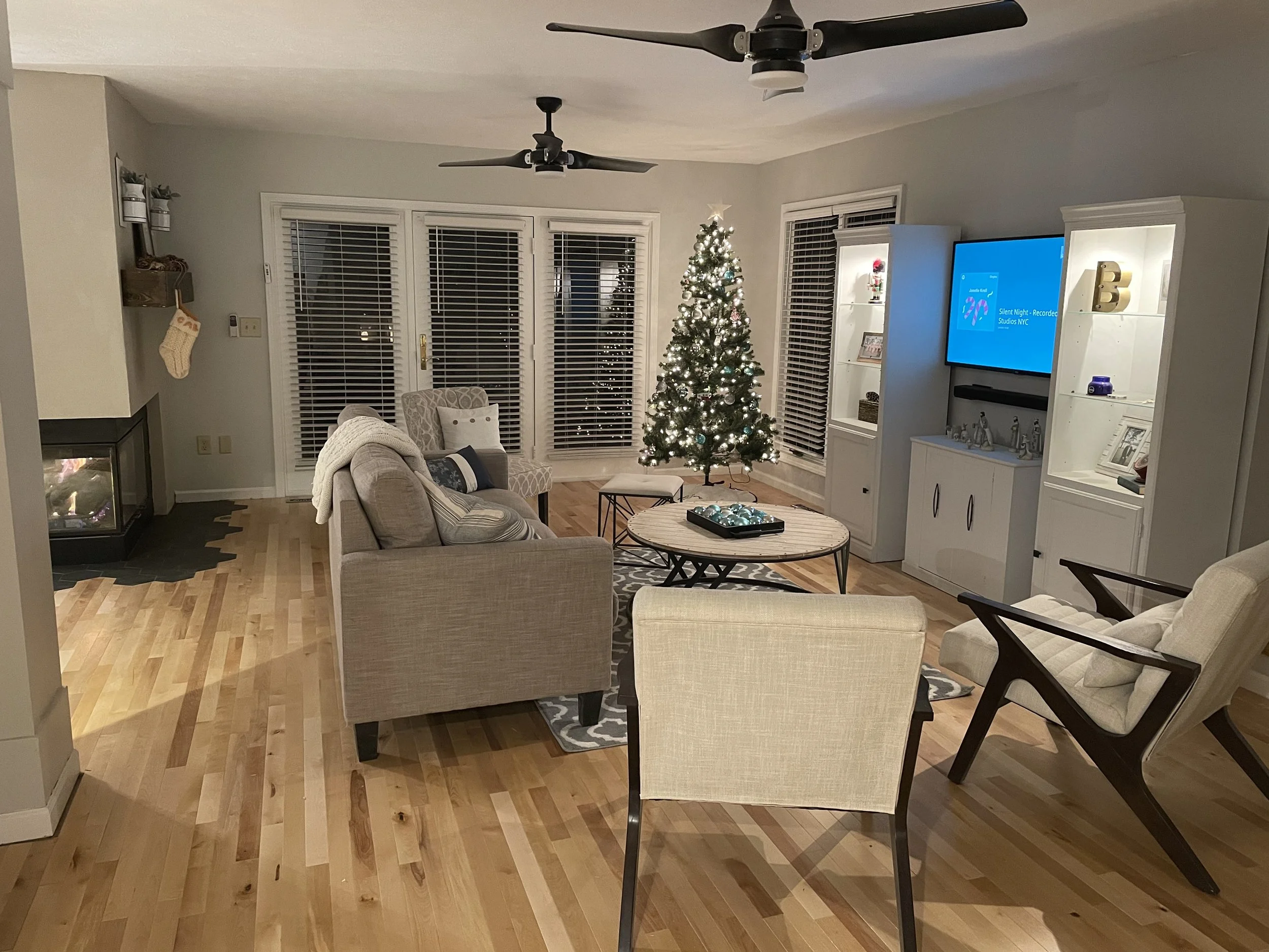 Living room decorated for Christmas with a lit Christmas tree, furniture including armchairs and sofas, a television on a white cabinet, and a fireplace on the left.