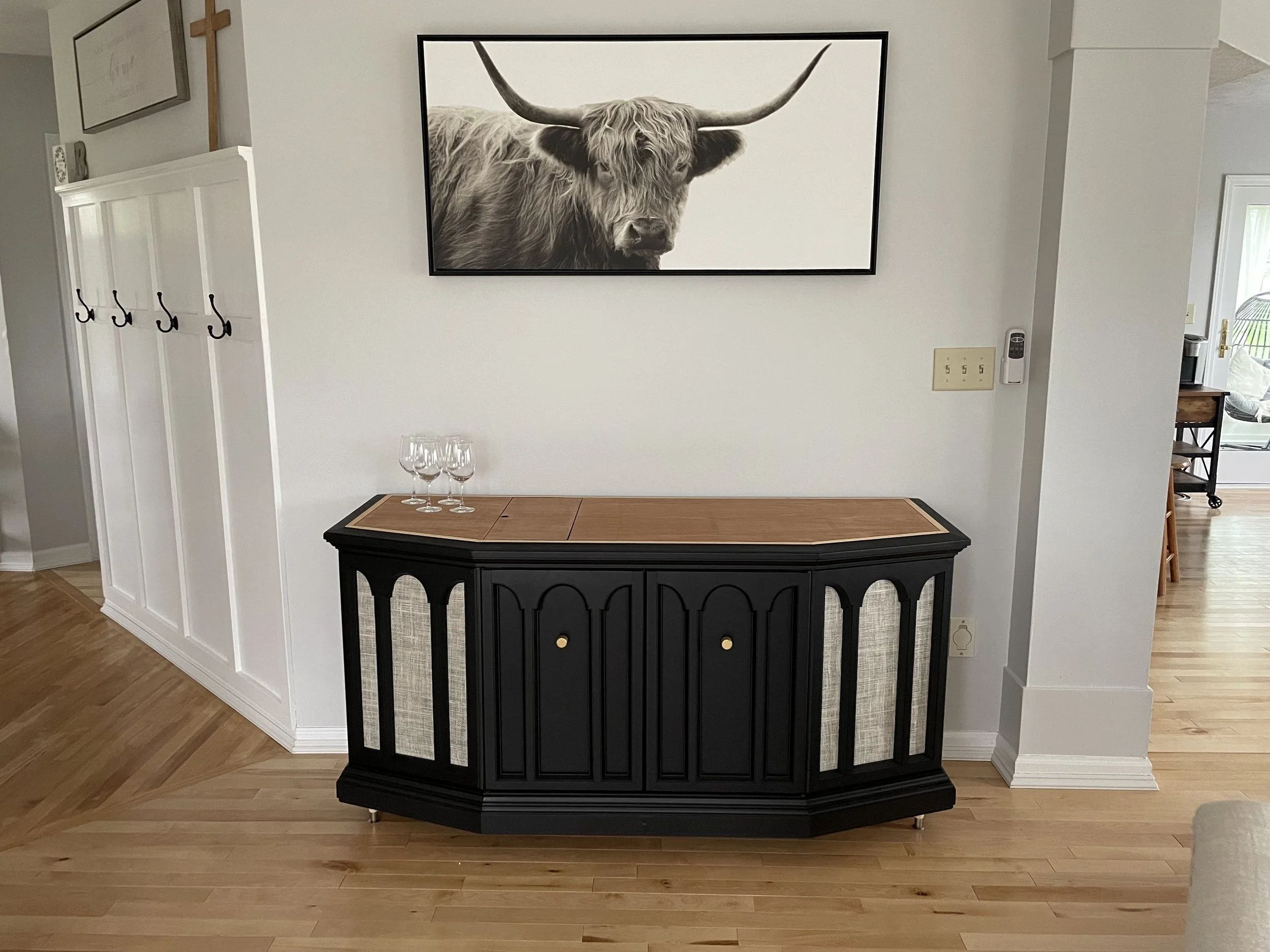 A black sideboard with a wooden top and textured panels on the doors, with three wine glasses on top, situated in front of a white wall with a large framed black and white photograph of a Highland cow above it.