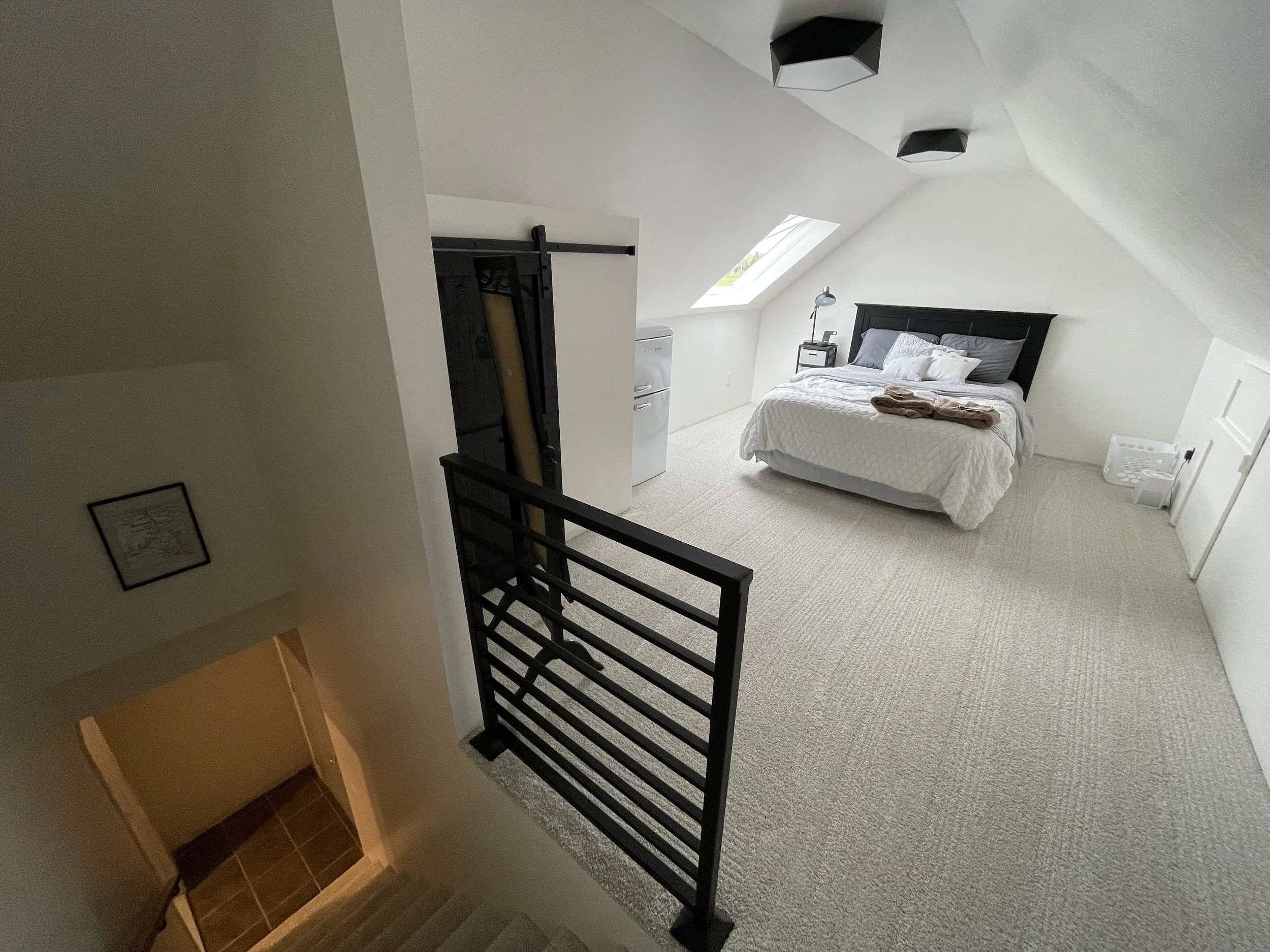 A bedroom with white walls and ceiling, a skylight window, a black bed frame with white bedding, a small nightstand with a lamp, and a laundry basket. There are folded towels on the bed and a sliding barn door near a small water heater.