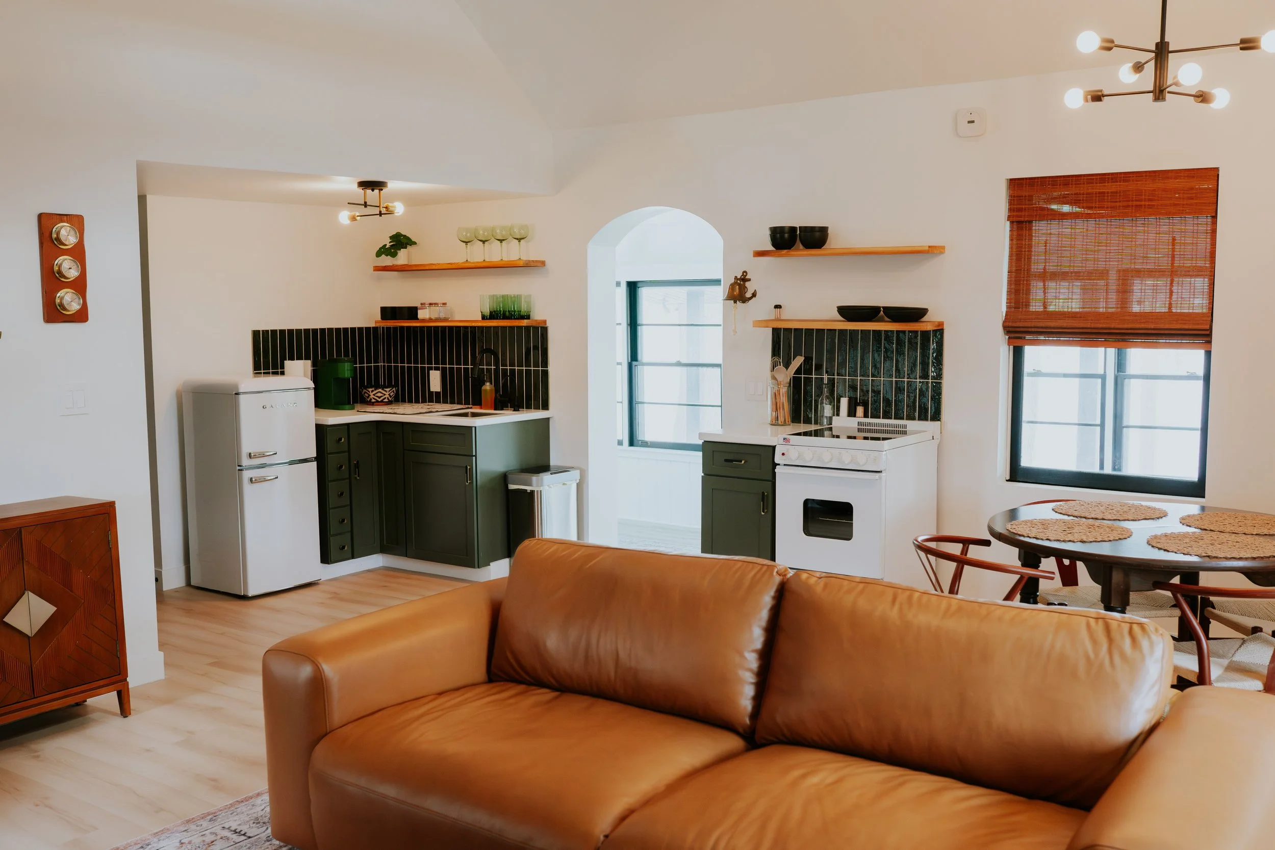 Open-concept living room and kitchen with white walls, green cabinetry, and black tile backsplash, featuring a tan leather sofa, dining table with chairs, and a window with blinds.