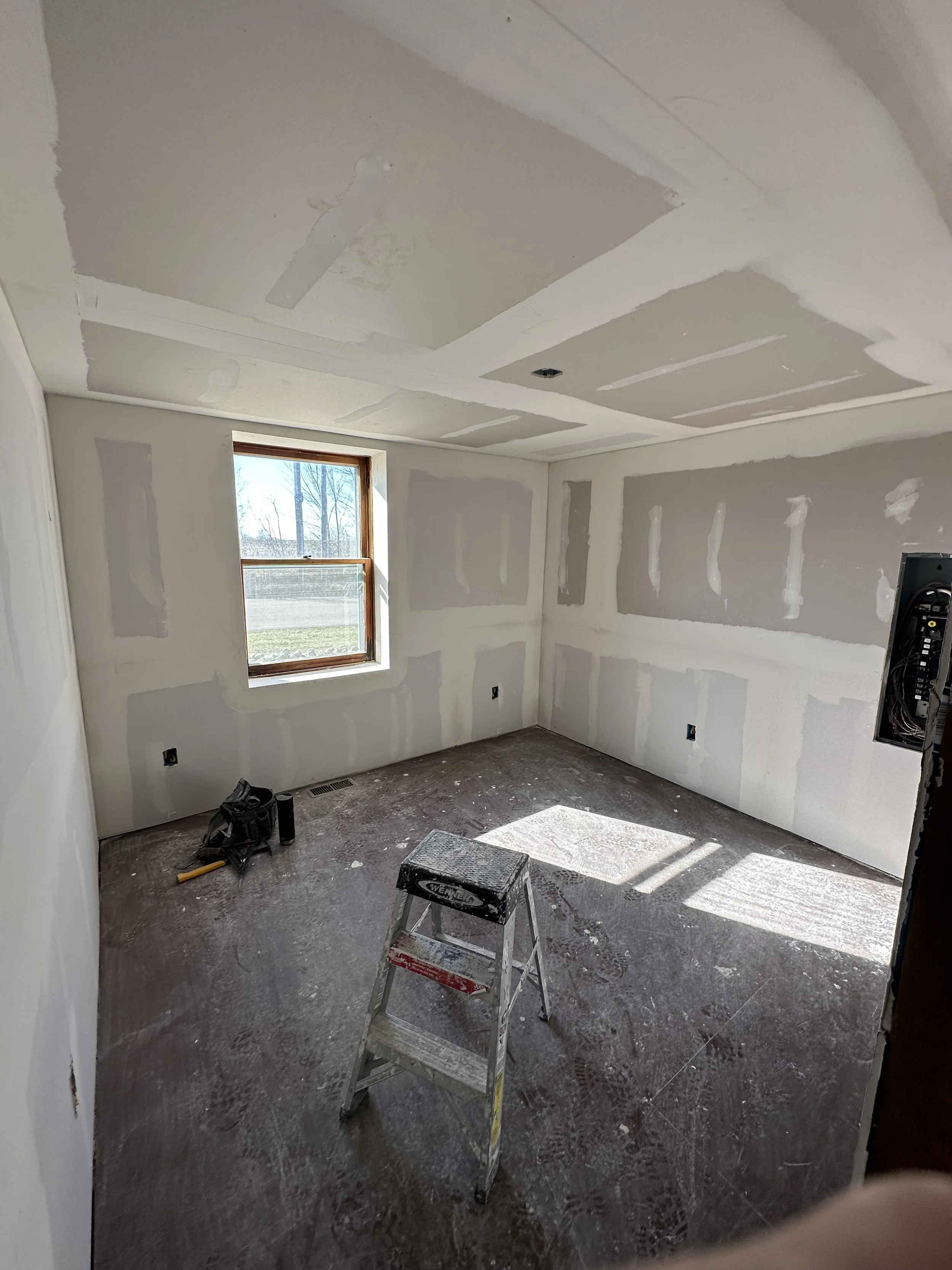 Empty room under construction with drywall and a small window, ladder and tools on the floor.