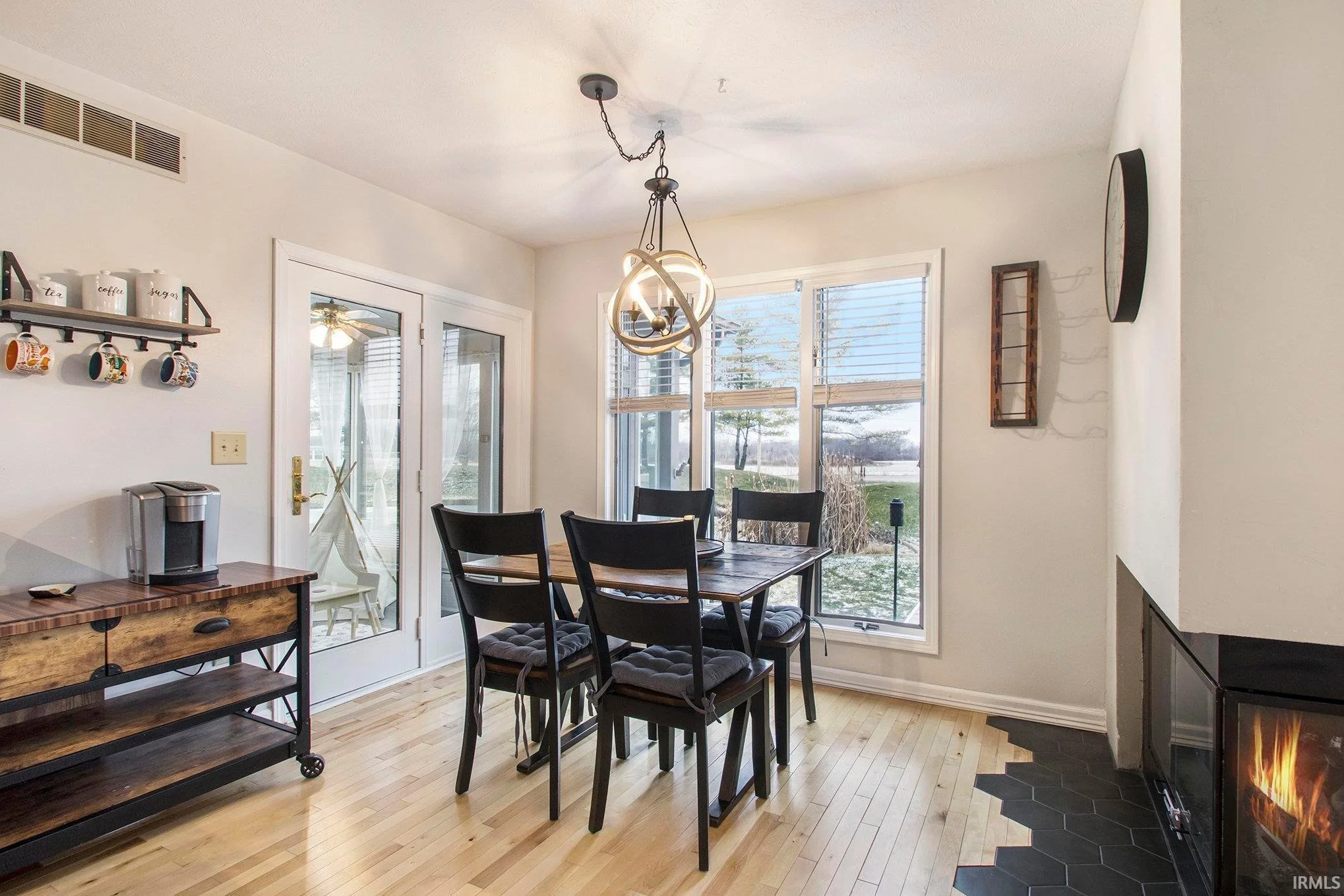 A dining room with a wooden table, six black chairs with cushions, a corner fireplace, and large windows showing an outdoor view with grass and trees. There's a decorative light fixture hanging from the ceiling, a small shelf with cups on the wall, and a small wooden cart with a coffee maker.