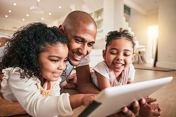 Family of three lying on the floor and smiling while looking at a tablet in a bright living room.