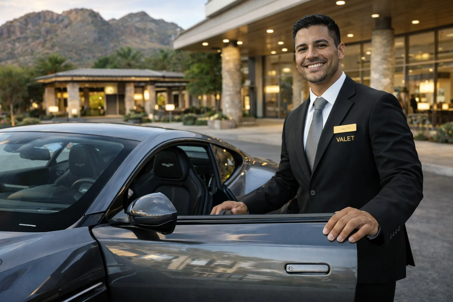 A valet attendant in a black suit, white shirt, and gray tie standing next to a gray sports car, smiling in front of a hotel with a mountainous background.