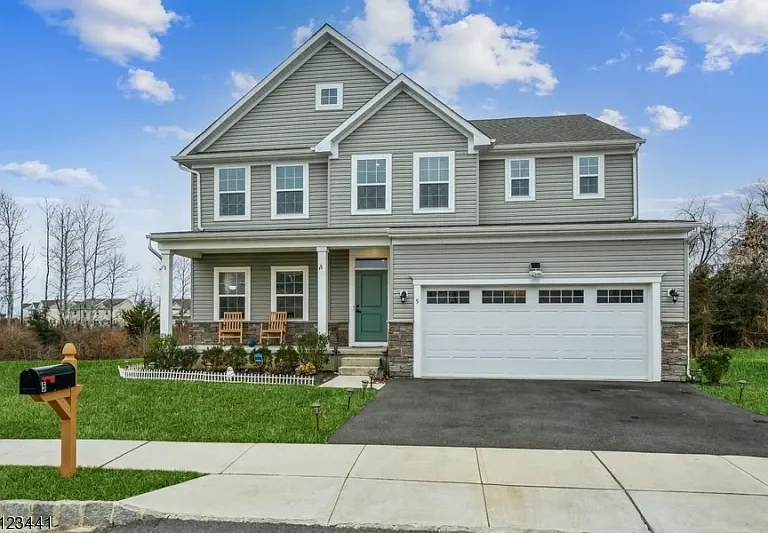 A two-story suburban house with gray siding, white trim, and a green front door. It has a two-car garage and a small front porch with two chairs. The front yard features a mailbox and a concrete sidewalk.