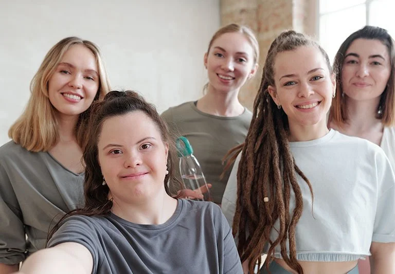 Five women smiling and taking a group selfie inside a bright room.