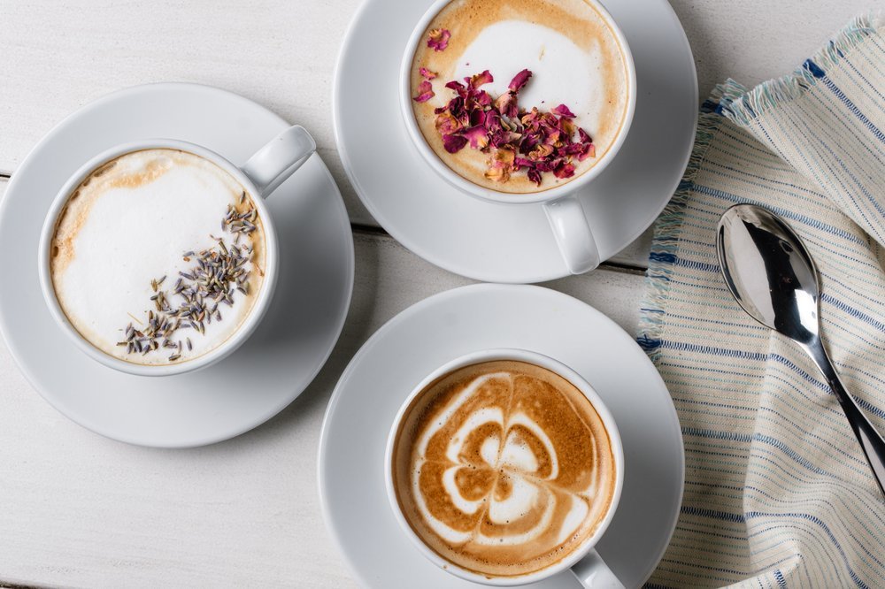 Three cups of coffee with different foam designs and toppings, placed on white saucers on a white wooden surface with a striped cloth and a silver spoon.