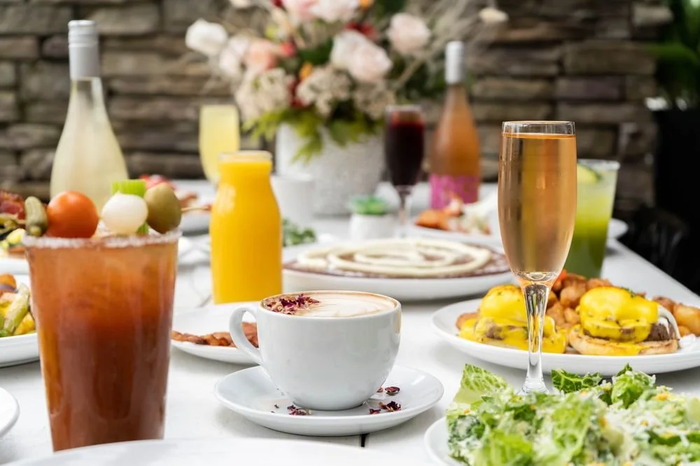 A table set with various dishes and drinks, including a salad, coffee, champagne, and bottles of juice or wine, with a background of flowers and a stone wall.