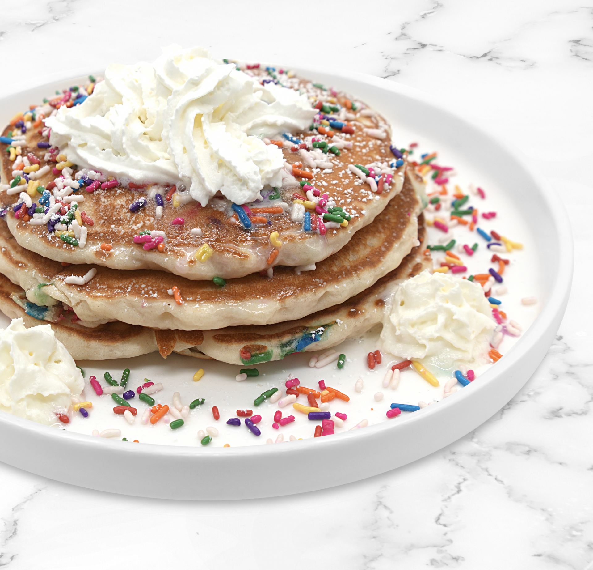 Three stacked pancakes topped with whipped cream, colorful sprinkles, and powdered sugar on a white plate.