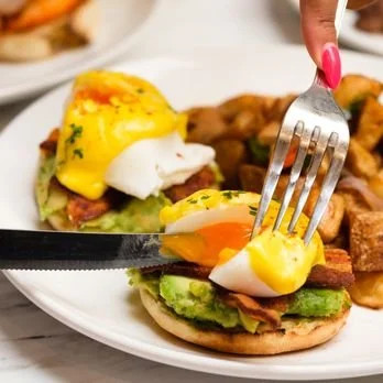 A plate with a breakfast sandwich topped with a poached egg and yellow sauce, alongside cooked meat and potatoes.