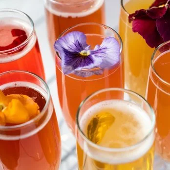 Variety of colorful drinks in tall glasses, some garnished with flower petals, on a white surface.