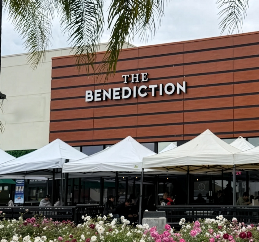 Exterior of a Benny's restaurant with outdoor seating and white tents, flowers in the foreground, and palm tree branches partially visible at the top.