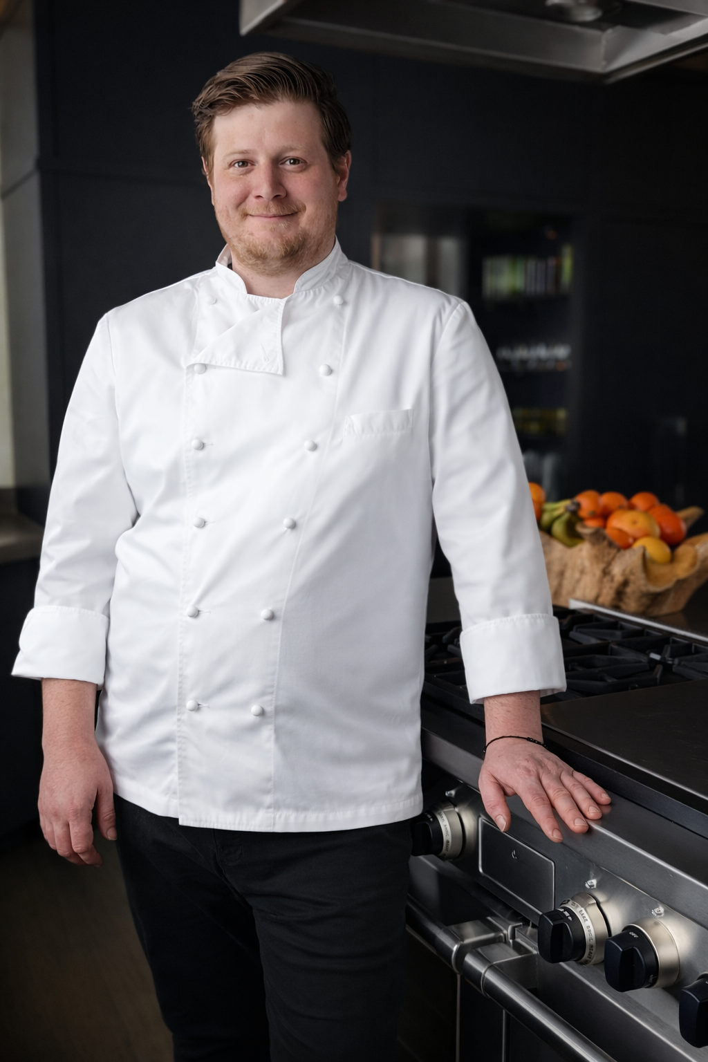 A male chef in a white chef's coat standing in a modern kitchen with a bowl of assorted fruits on the counter.