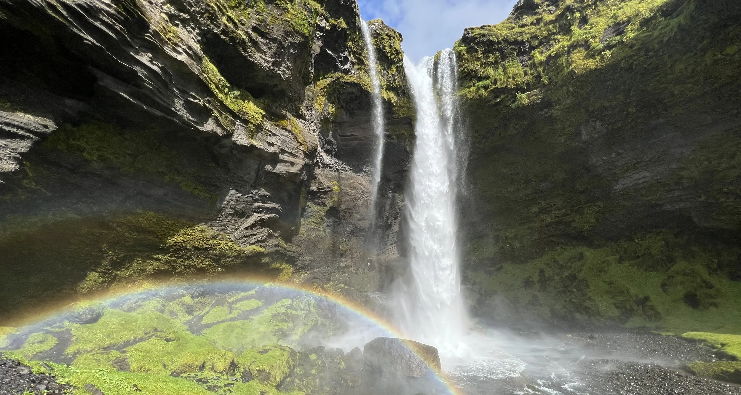 A waterfall cascading down between moss-covered rocks, with mist and a rainbow at the base of the falls.