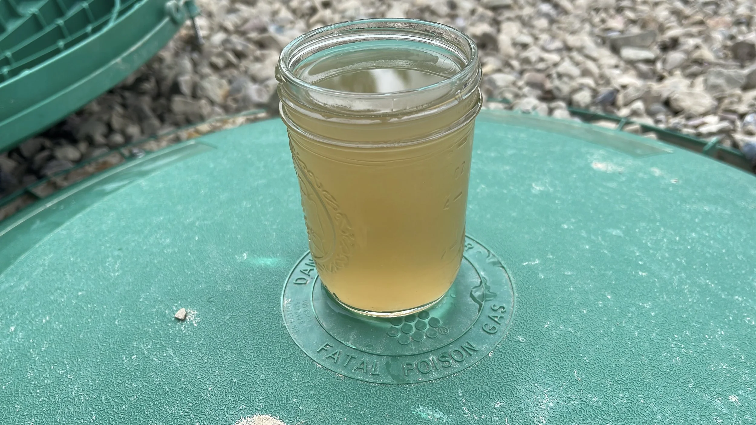 A glass jar filled with a cloudy, brown liquid is placed on a green surface, outdoors on a gravel ground.