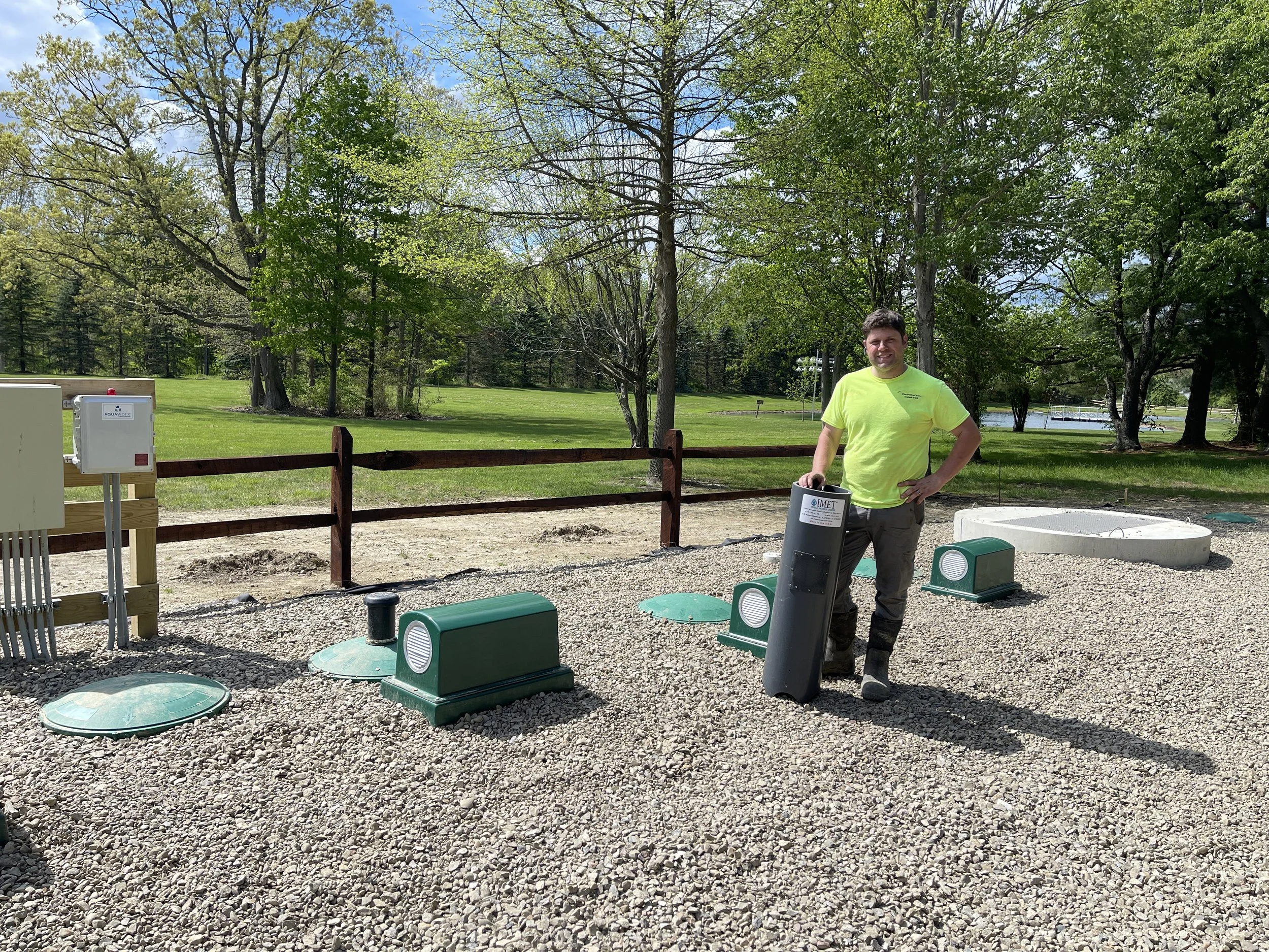 A man standing outdoors in a park or recreational area, wearing a bright yellow shirt and dark pants, posing next to some green and gray IMET modules and pump gousing on gravel ground with trees and grass in the background.