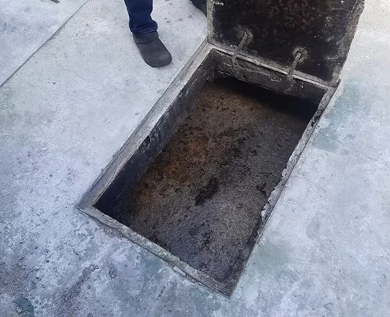 Open manhole on a concrete sidewalk with a person standing nearby. The manhole cover is raised, revealing a dark underground opening and heavy buildup.