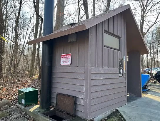 Small wood structure with a slanted roof, located in a wooded area, with a window, a sign, electrical meters, and a trash bin nearby.
