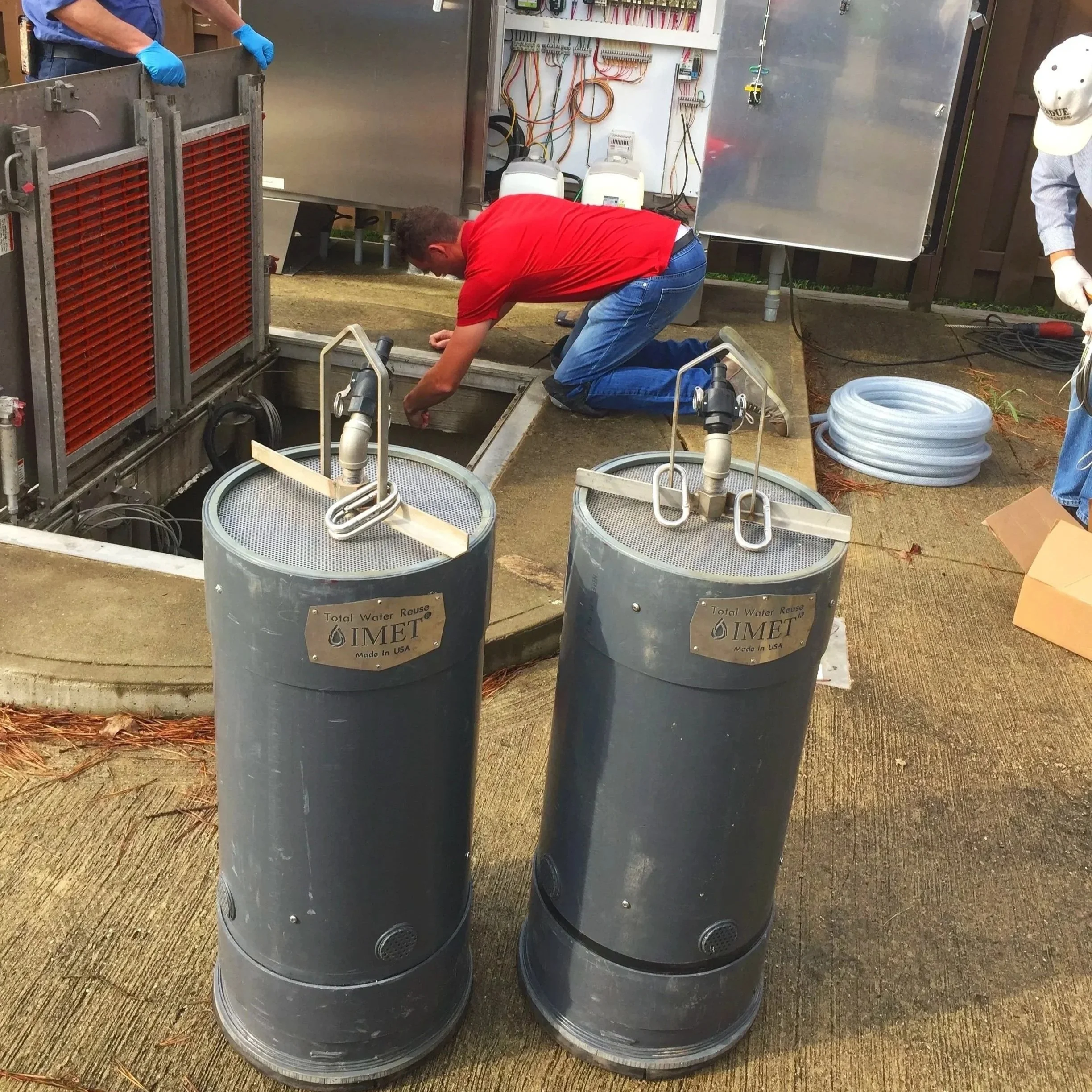 Two large gray water treatment modules labeled 'IMET Total Water Reuse' in front of a man in a red shirt and blue jeans, who is working on an underground tank, with two other people partially visible in the background.