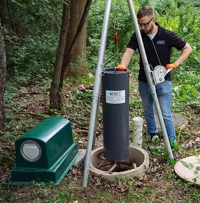 A man installing a device in a wooded outdoor area, with a large green equipment box nearby and a black cylindrical wastewater treatment module hanging from a tripod.