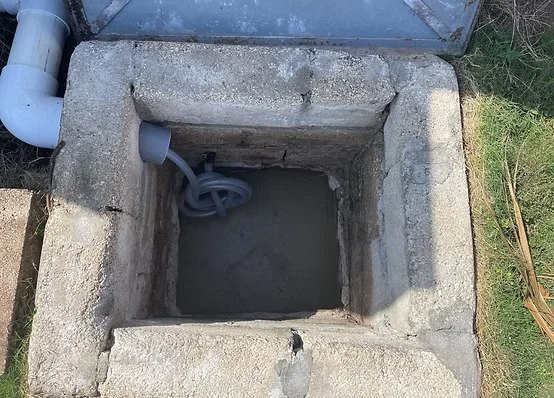 An outdoor concrete well or pit with a PVC pipe and hose inside, located next to a metal fence and some grass. Heavy buildup.