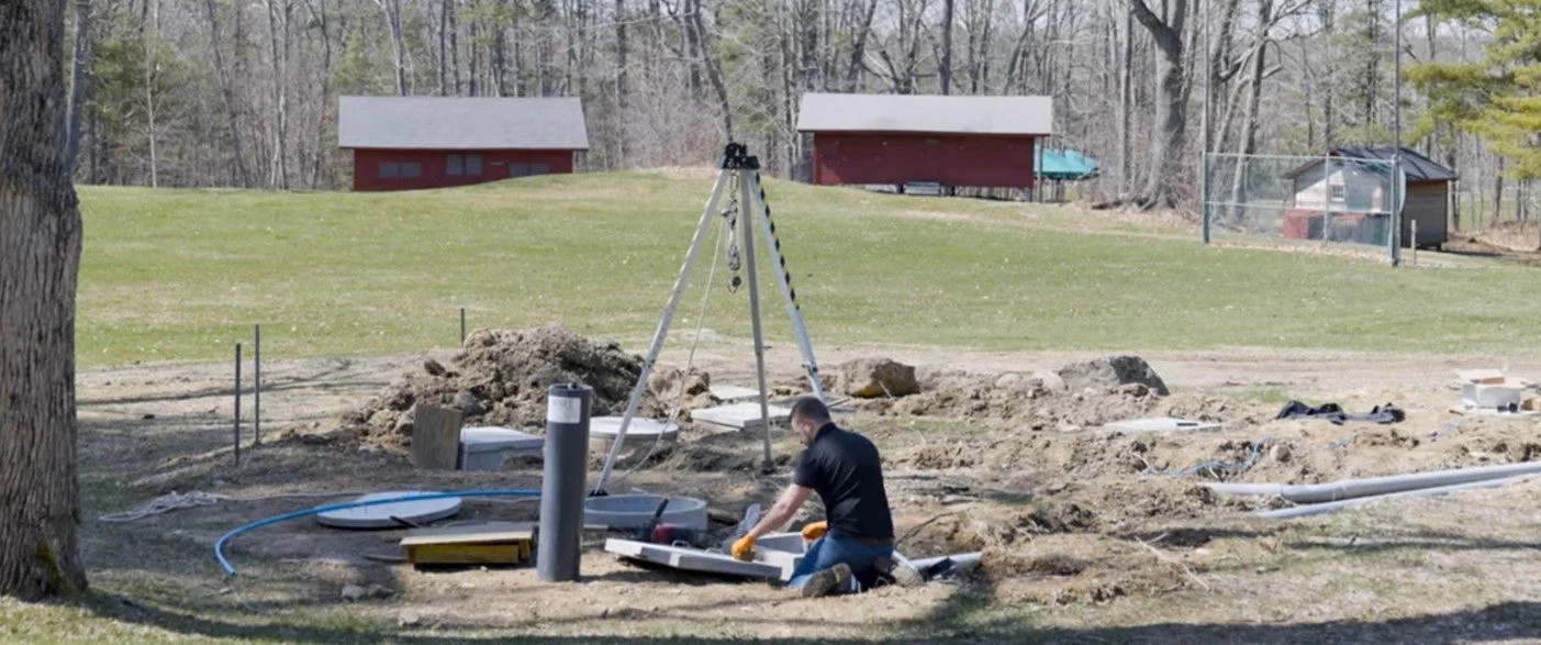 A man working on installation or maintenance of underground utility pipes in a backyard or park with trees and two red sheds in the background.