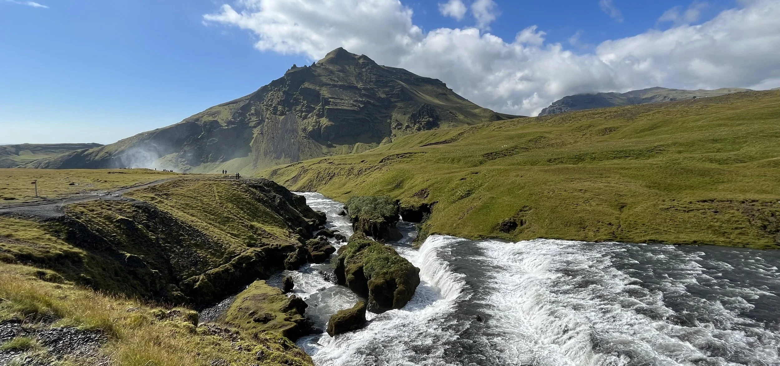 A green mountain with a stream flowing through grassy terrain under a partly cloudy sky.