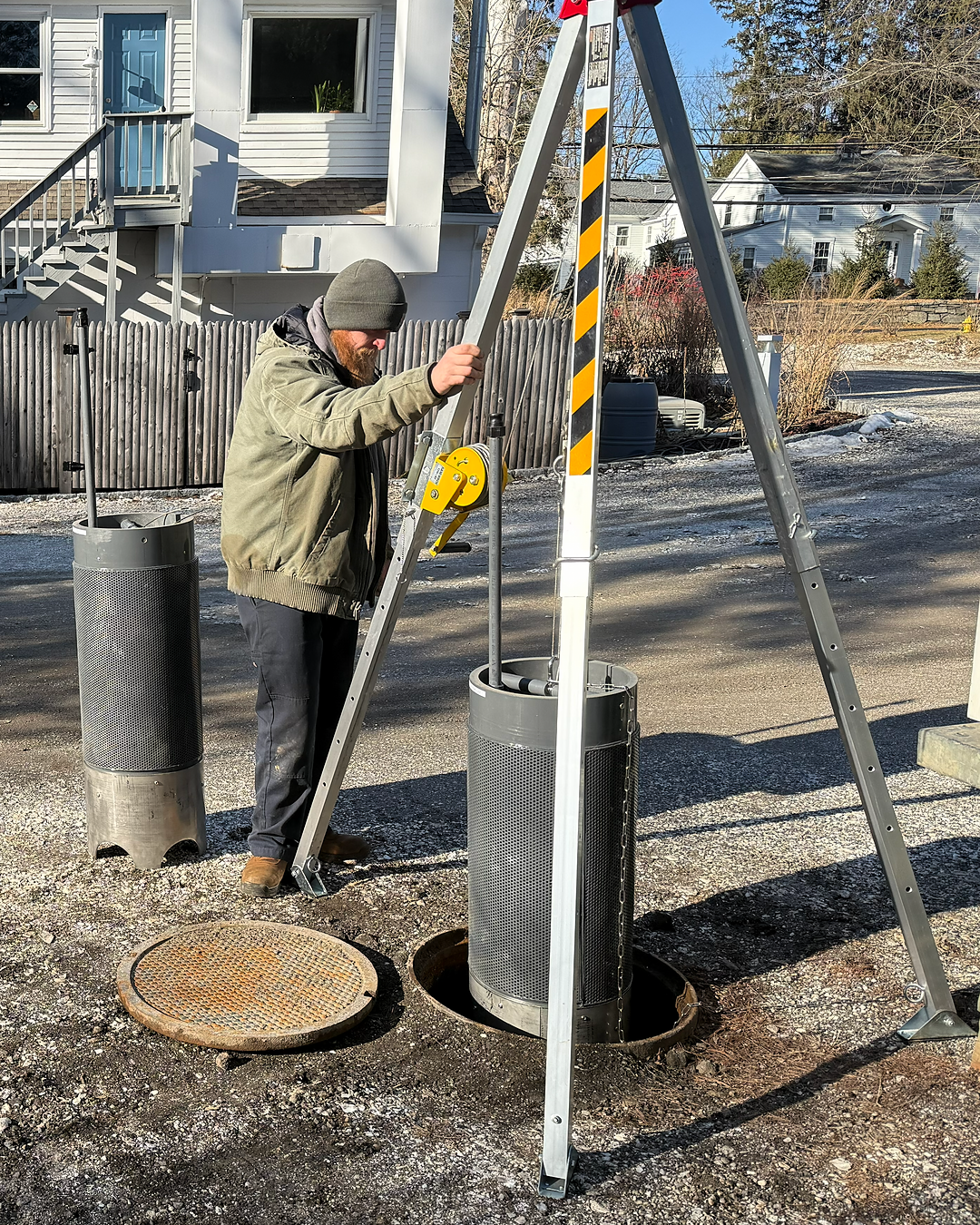 A man wearing a gray beanie and green jacket installing an IMET Grease Module, standing on a ladder next to a manhole with a round cover.
