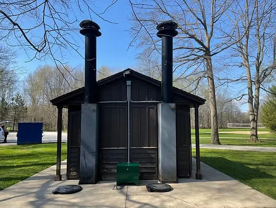 A small black wooden building with two large black pipes on top, situated on a concrete pad in a park with trees and a green grassy area in the background.