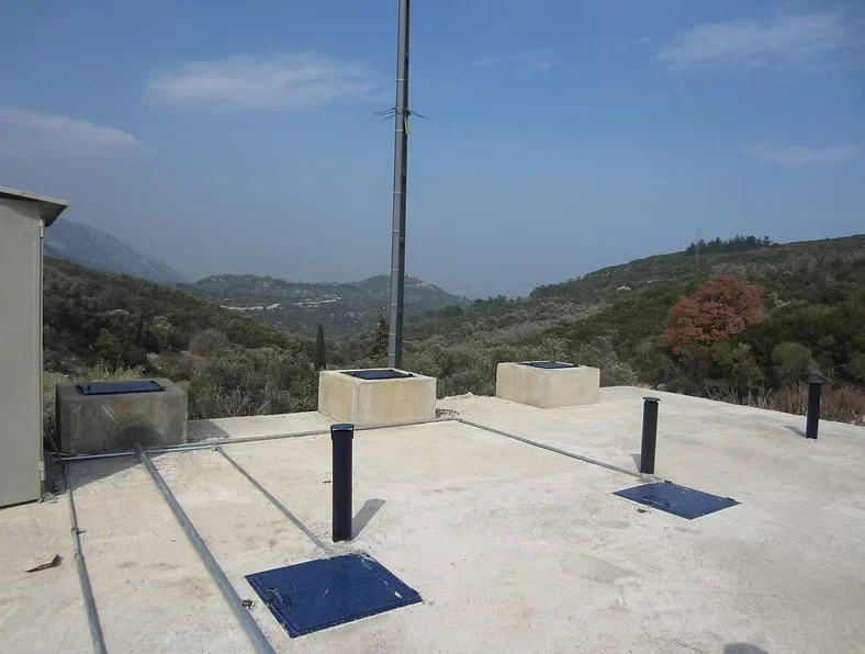 View from a wastewater treatment plant with two vent covers on the concrete surface, overlooking a hilly landscape with trees and mountains.