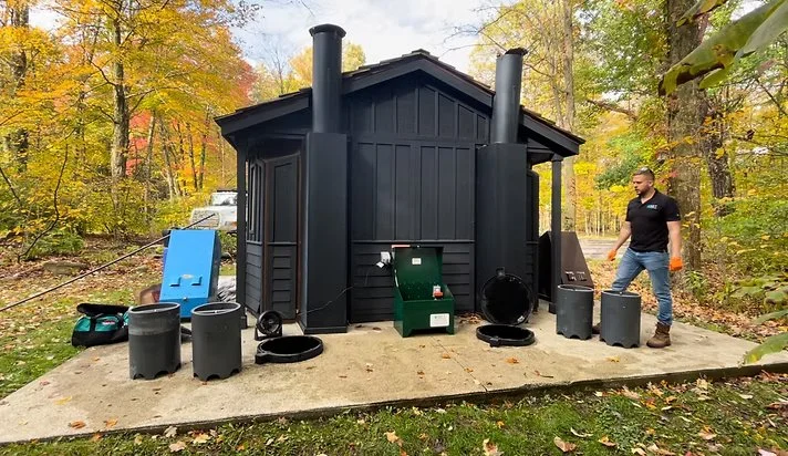 A man working on camo vault toilet, surrounded by trees with autumn foliage. IMET modules and tools are nearby.