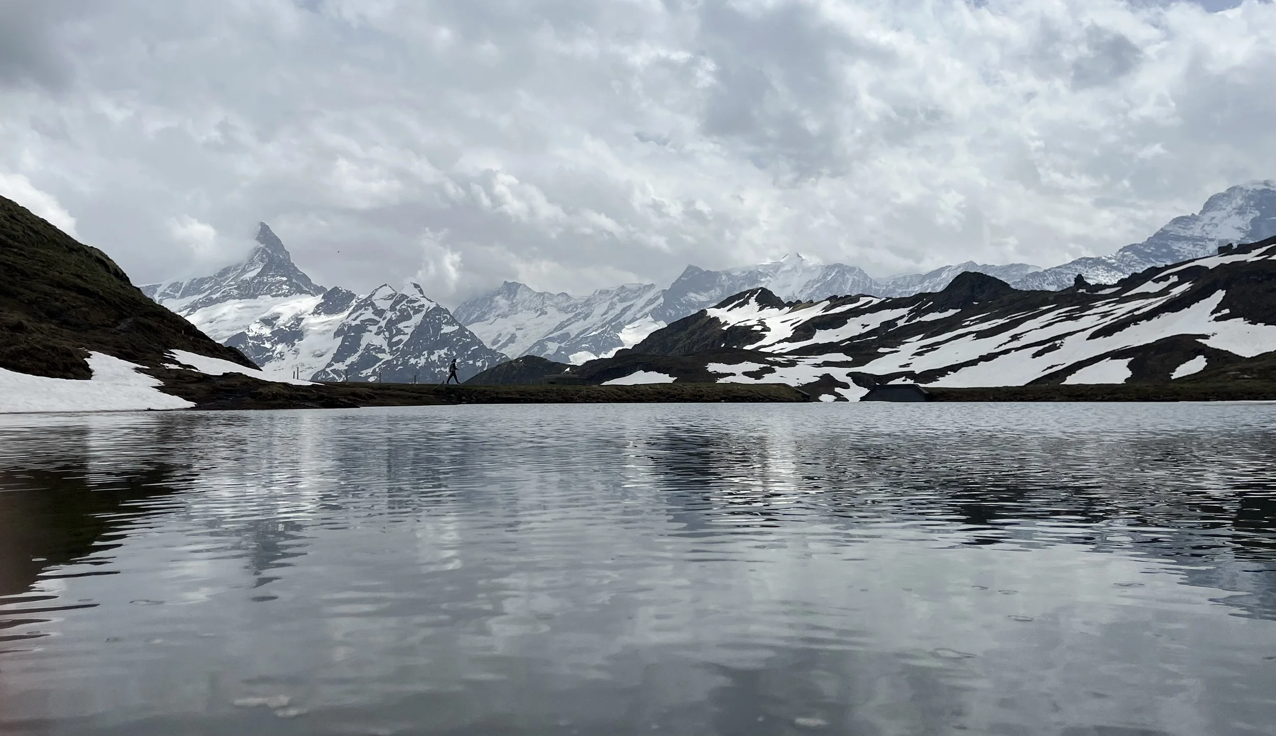 Snow-capped mountains surround a reflective lake under a cloudy sky, with a person walking along the shoreline.