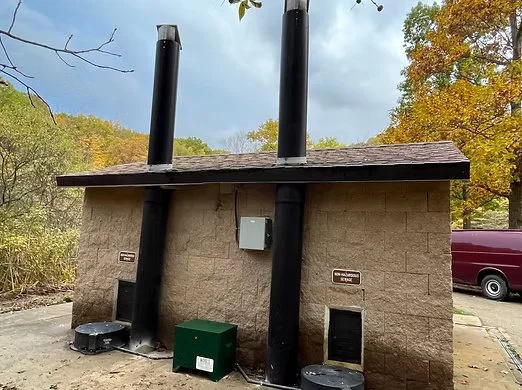 Back of a small vault toilet building with two large black exhaust pipes extending from the roof, surrounded by trees with fall foliage.