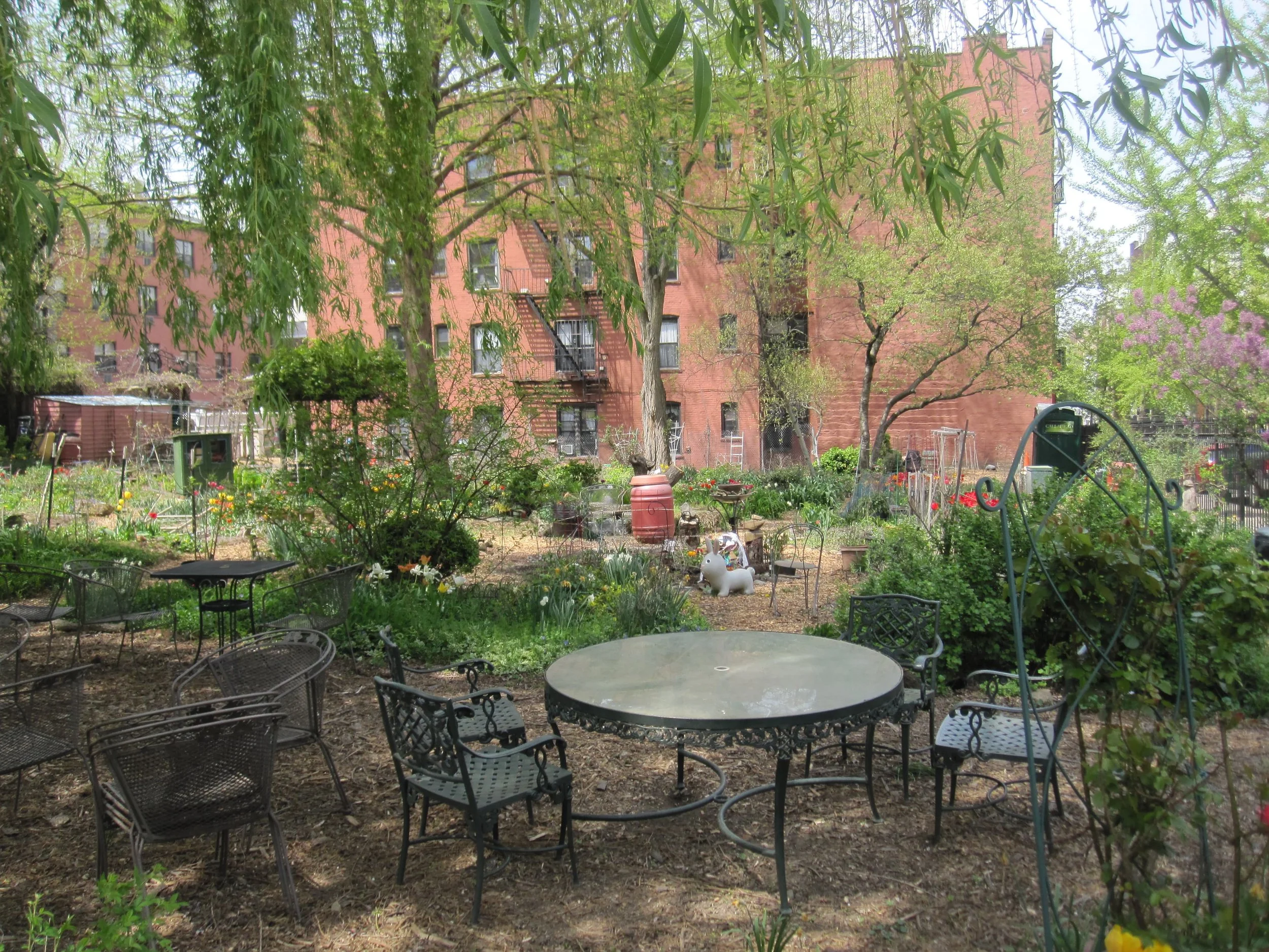 A Brooklyn urban garden with outdoor metal tables and chairs, surrounded by trees and flowering plants, with a brick apartment building in the background.