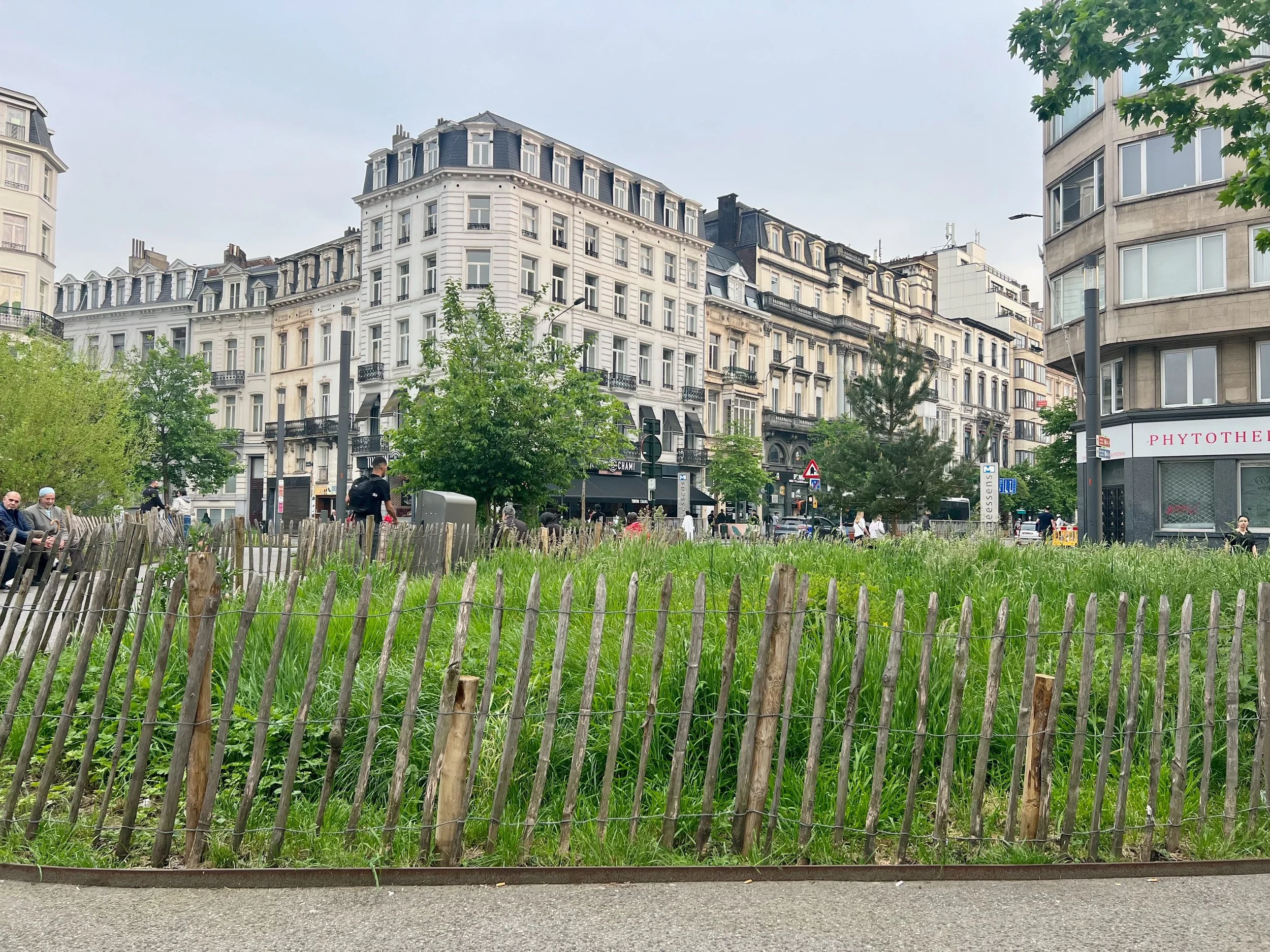 Brussels urban landscaping with a wooden fence, green grass, trees, and people sitting on benches or walking. Multi-story buildings line the street in the background.