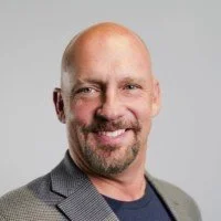 Bald man with a beard smiling, wearing a gray blazer and dark shirt