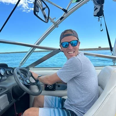 Smiling man wearing sunglasses and a cap, sitting at the steering wheel of a boat on a sunny day with blue skies and water in the background.