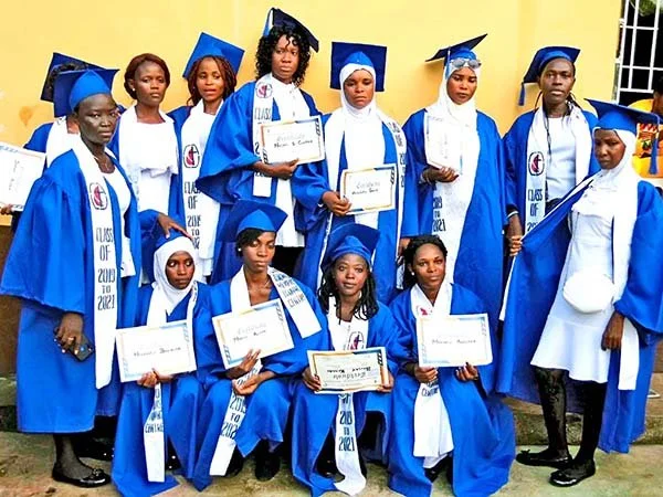 Group of graduating students dressed in blue caps and gowns holding certificates, standing outdoors in front of a yellow wall.