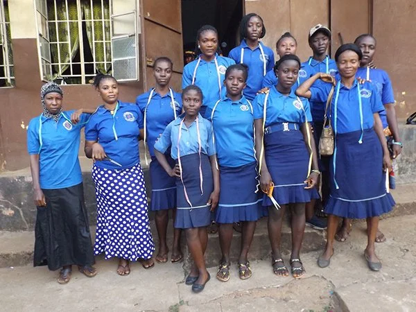 Group of women and girls in matching blue shirts and skirts standing outside a building.