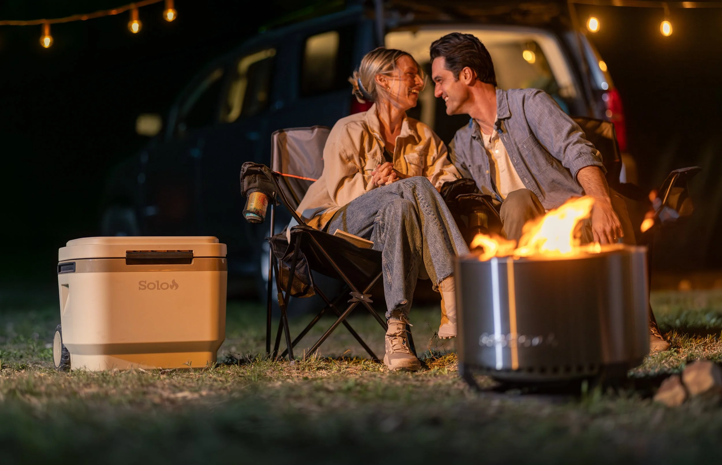 A smiling couple sitting near a campfire at night, enjoying each other's company with a parked car and string lights in the background.