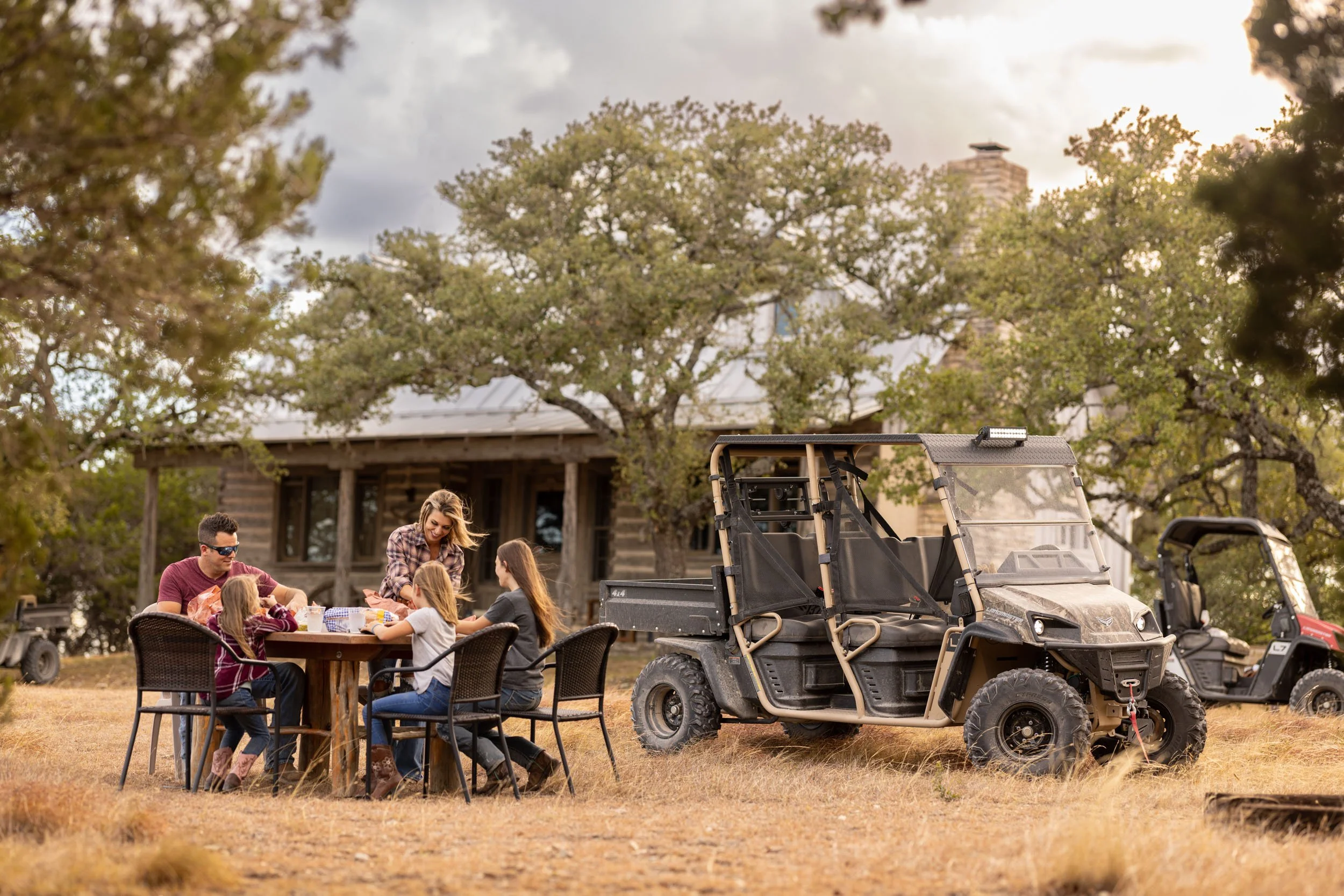 People gathered around a table outdoors with trees and a house in the background, and utility vehicles parked nearby.