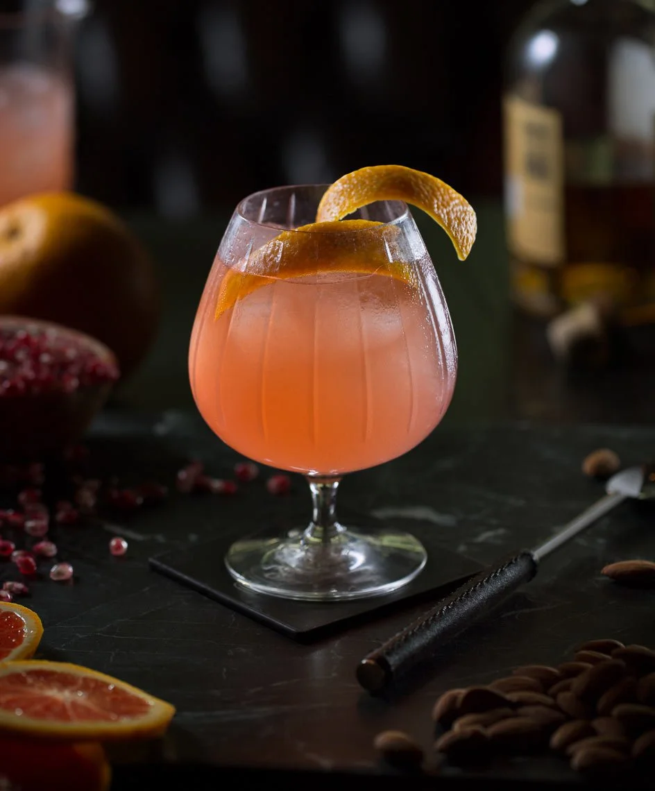 A pink cocktail with lemon twist garnish in a stemmed glass on a dark surface with pomegranate and blood orange slices and scattered nuts around.