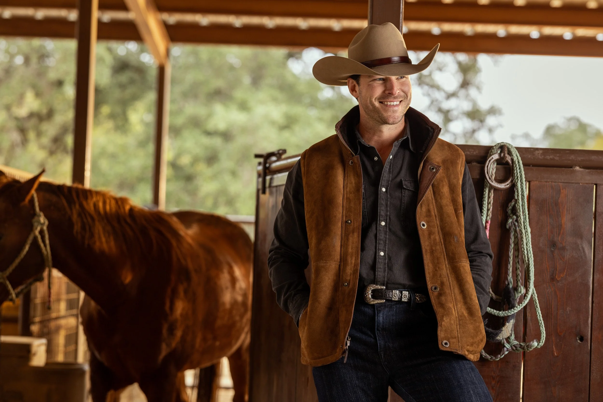 Smiling man in cowboy hat and brown vest standing in a horse stable with a brown horse nearby.