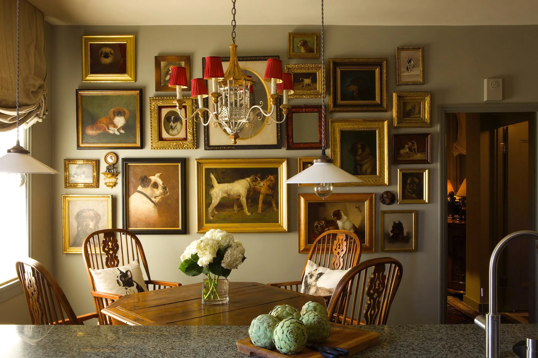 A cozy dining room with a wooden table and chairs, adorned with a vase of white hydrangeas. The wall features numerous framed paintings and photos of dogs, primarily in portrait style, with some in ornate gilded frames and others in simple wood or bl