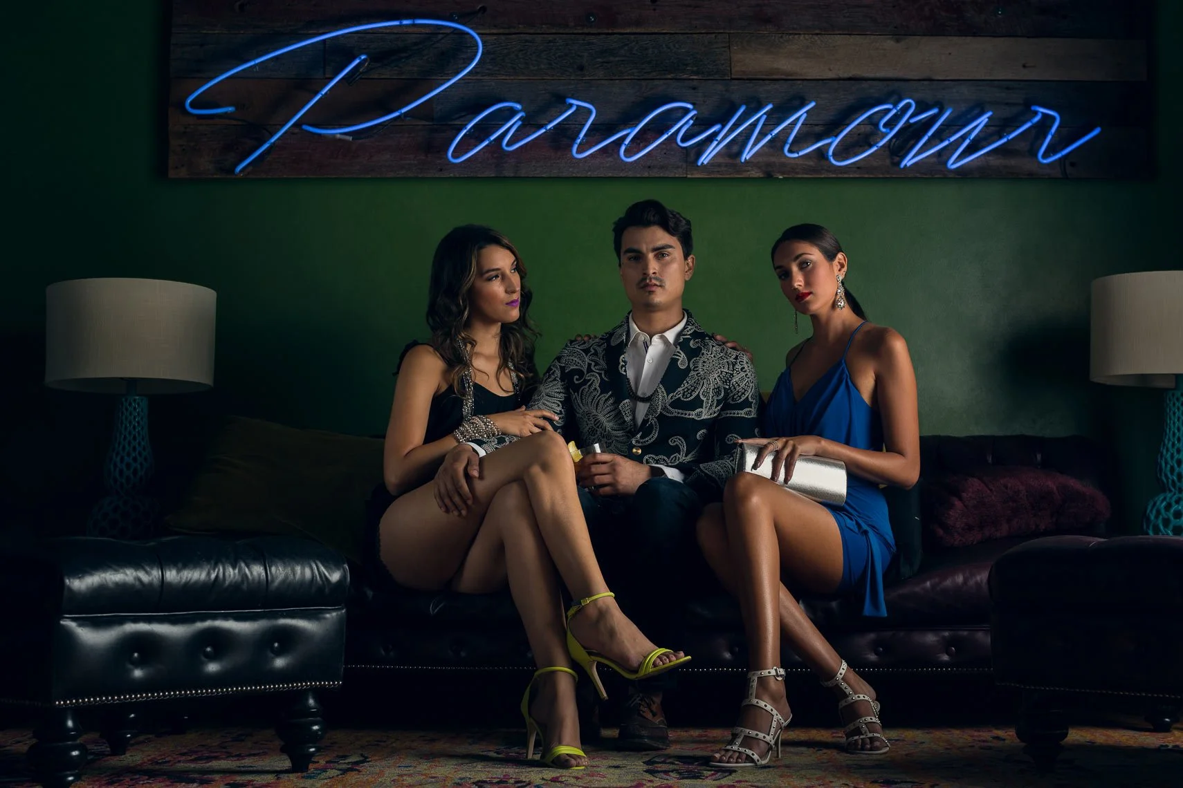 Three people sitting on a black leather sofa in front of a green wall with a blue neon sign that says 'Paradise,' with two lamps on either side of the sofa. The individuals are dressed stylishly, with the women in cocktail dresses and high heels, and