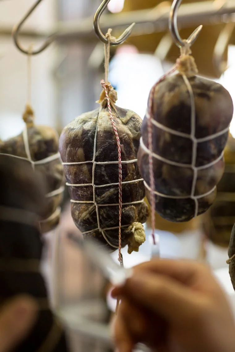 Close-up of cured meat hanging in a shop, wrapped in white netting with twine at the top, hanging from metal hooks.