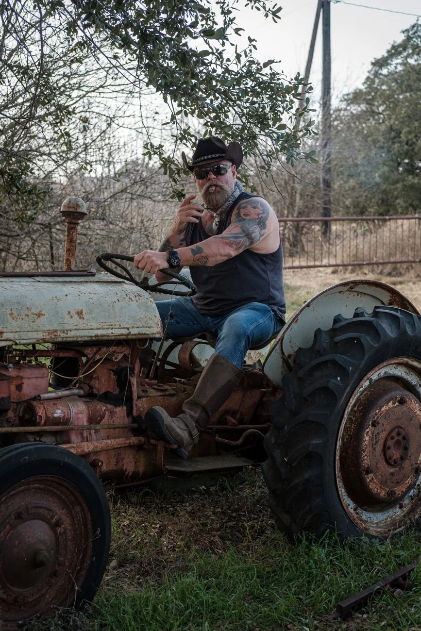 A man with tattoos, sunglasses, a cowboy hat, and a cigar sitting on an old, rusty tractor outdoors surrounded by trees and grass.