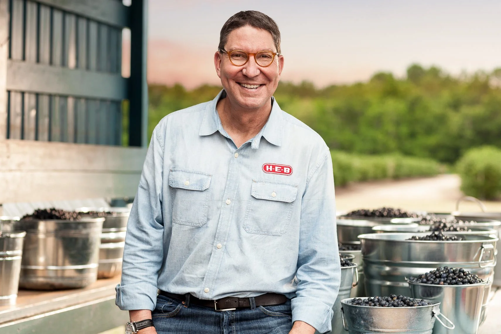A smiling man wearing glasses and a light blue button-up shirt with an H-E-B logo, standing outside near metal buckets filled with blueberries, with a backdrop of green trees and an orange-pink sky.