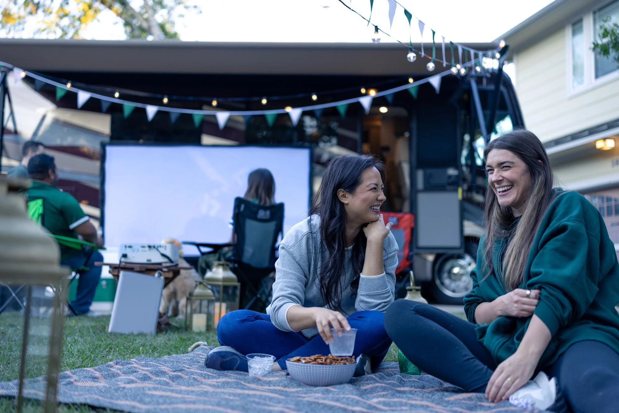 Two women sitting on a blanket in a backyard, enjoying food and drinks, with a mobile movie screen and string lights behind them, indicating a casual outdoor gathering.
